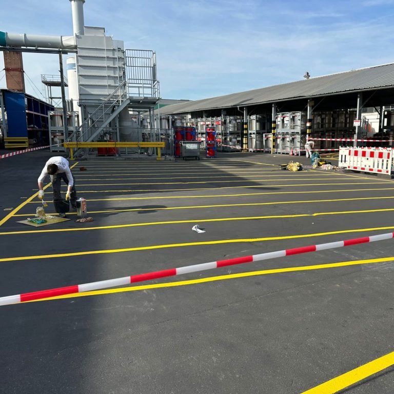 Mann markiert gelbe Linien auf Industriegelände mit Industriegebäuden im Hintergrund.